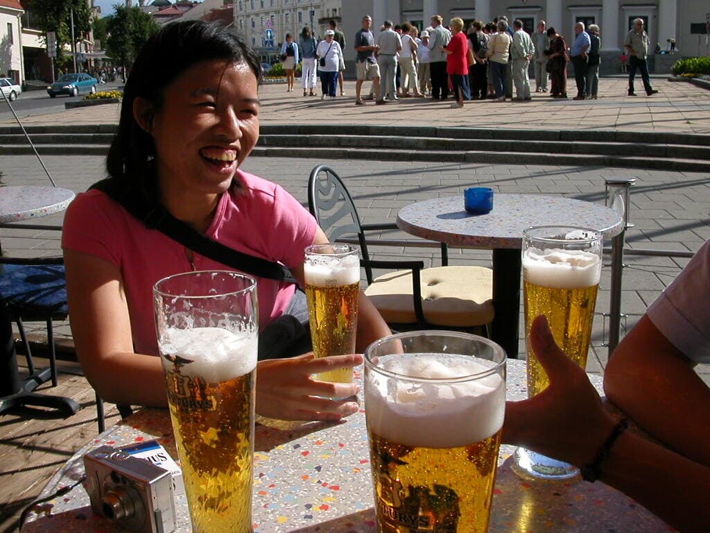 One of us laughing over tall glasses of beer on a café terrace in Lithuania, a crowd gathering in the square behind