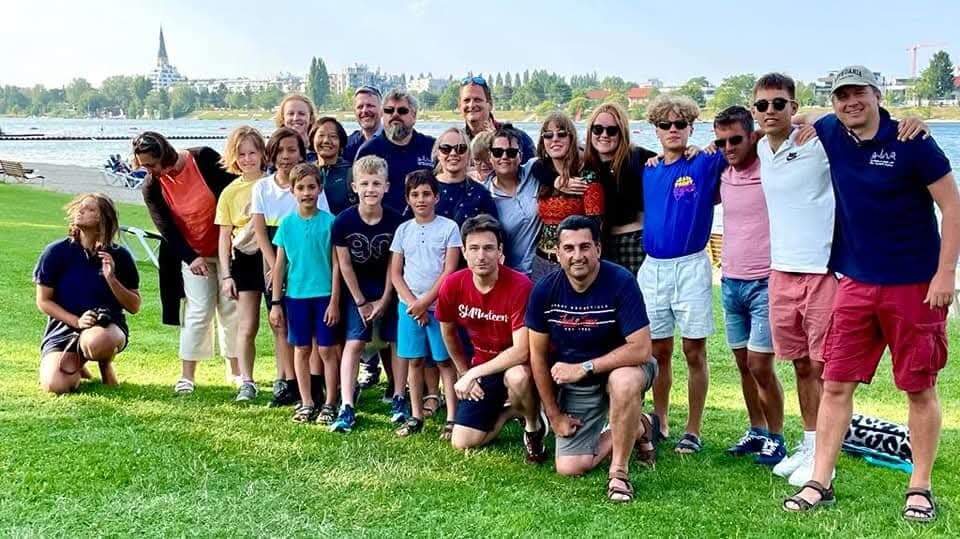 The whole gang, kids included, on a lakeside lawn, a church spire across the water