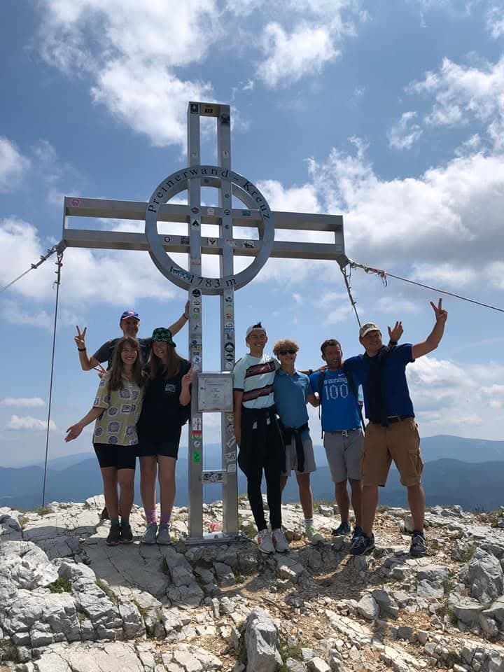 Eight of us at a summit cross on a rocky mountain top, arms in the air