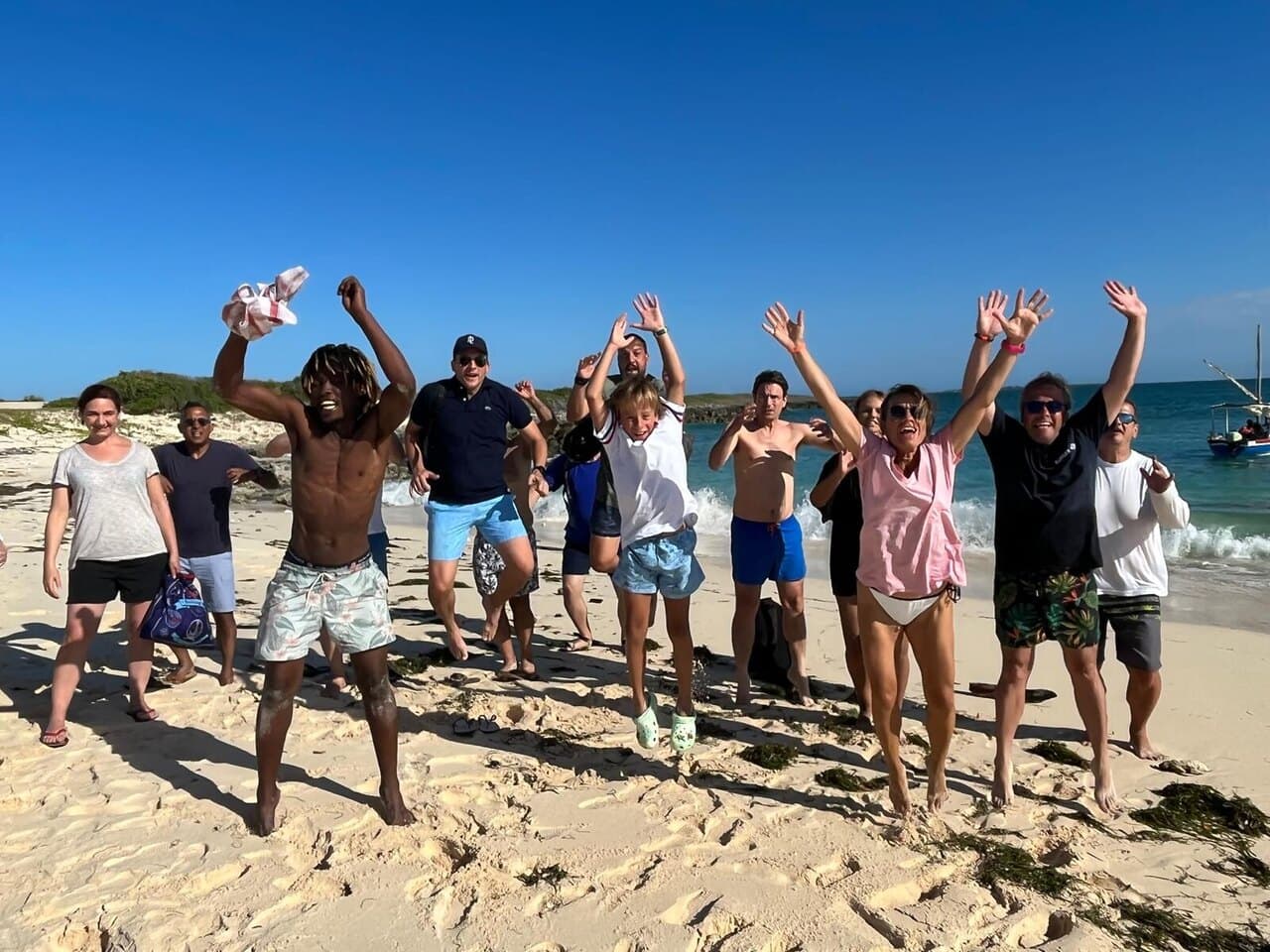 A dozen of us jumping and cheering on a white beach, a dhow moored behind