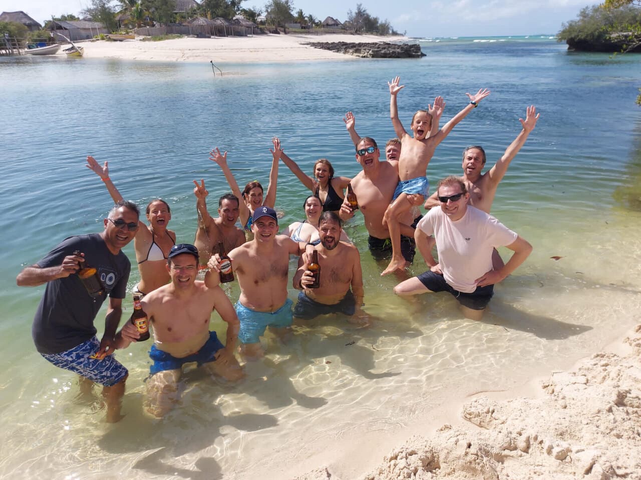 A crowd of us cheering knee-deep in clear water off a white beach, a child up on someone's shoulders