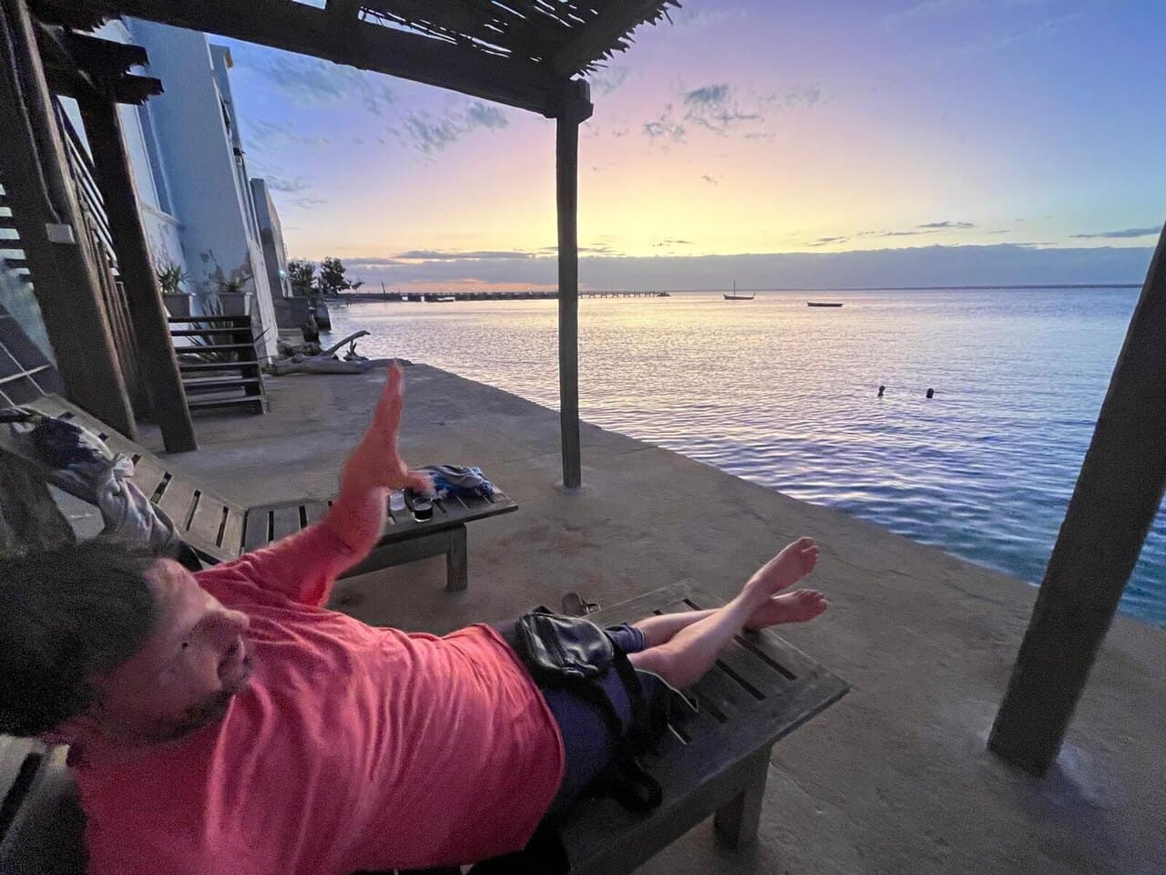 One of us stretched out on a deck chair, waving, as the sun sets over the sea